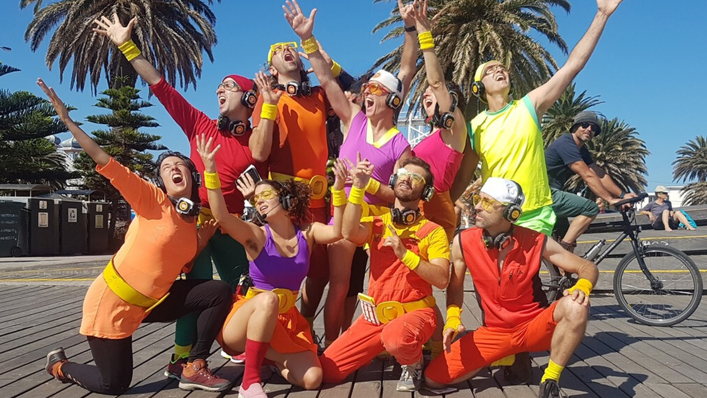 Nine people standing in a group posed. they are all wearing heaphones and colourful bright clothes. they look happy and are singing. 