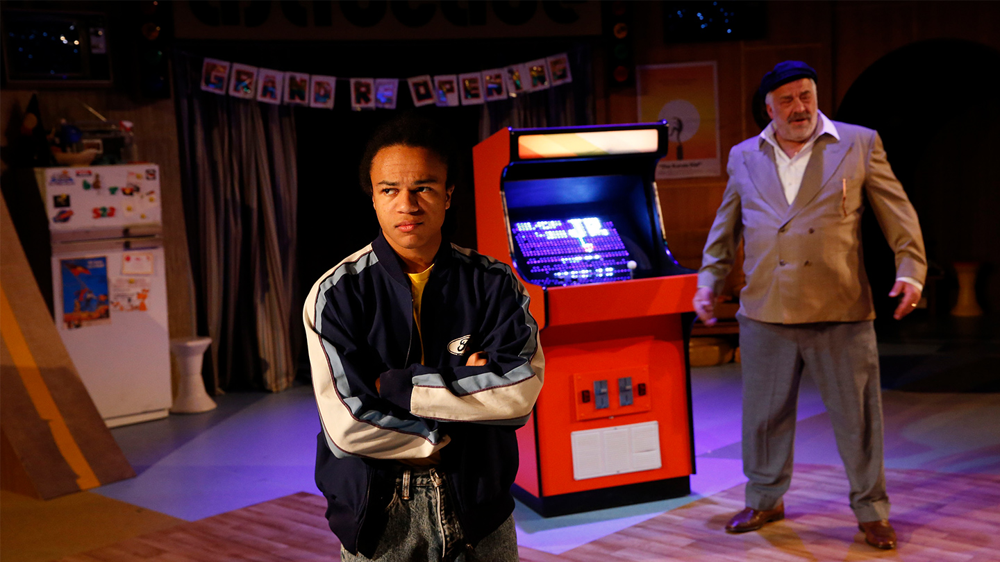 A production image from the Melbourne Theatre Company's season of Astroman, showing a man in foreground wearing a white tshirt and tracksuit top who has crossed his arms. In the background there's an arcade machine and an older man standing next to it.
