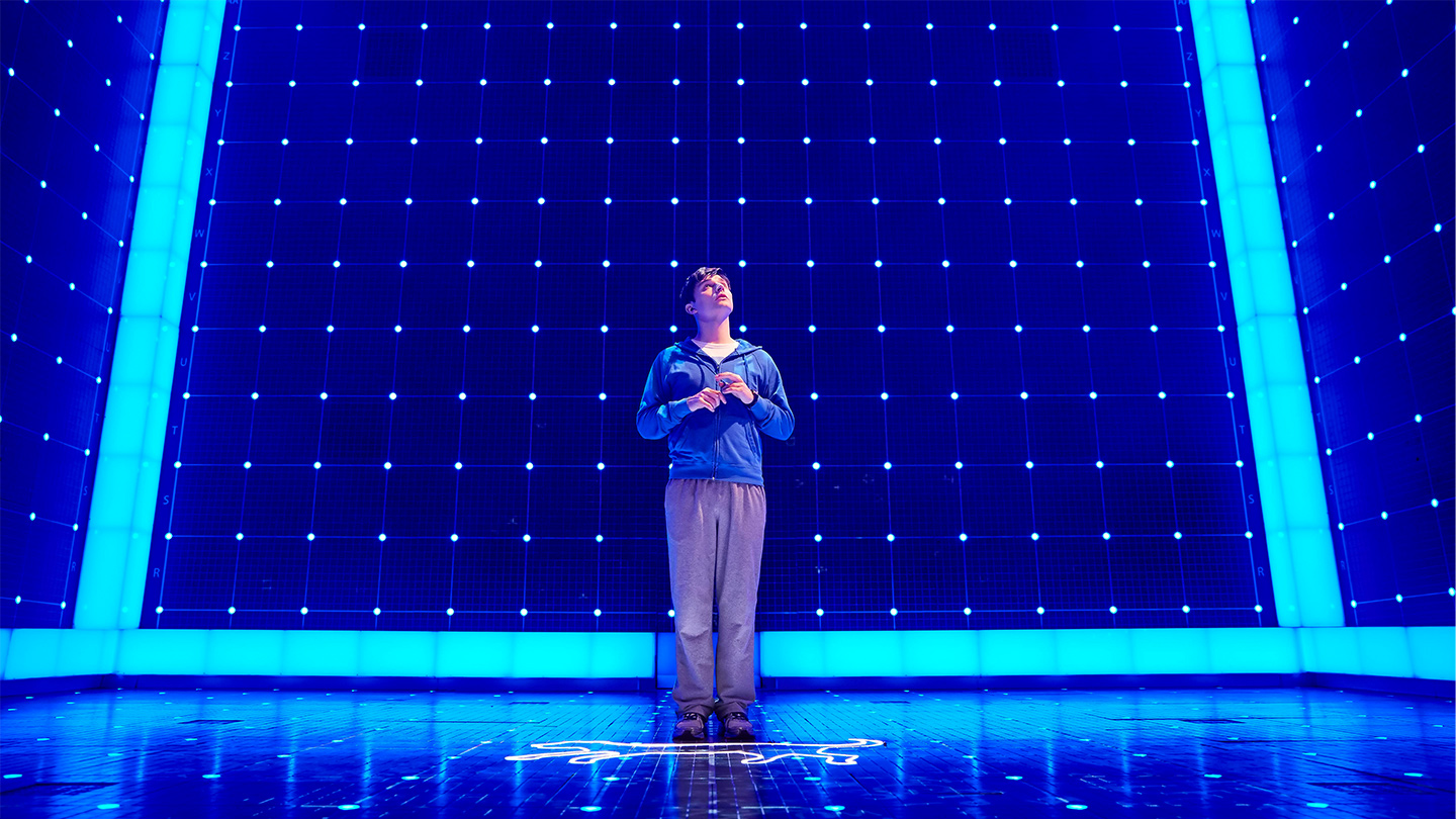 A production image of the Melbourne Theatre Company's season of The Curious Incident of the Dog in the Night-Time showing a young man standing in the centre of a stage wearing a blue jumper and tan trousers, his hands held up to his chest. Behind him, blue neon frames the edges of the stage.