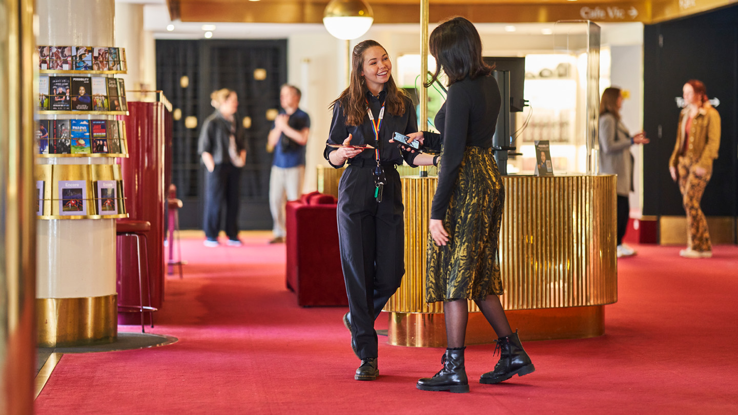  A lady is being served by an Arts Centre Melbourne staff member. They are standing in the foyer of the Arts Centre. The staff member is wearing black pants and a black shirt. The lady being served is facing her, wearing a yellow skirt and black jumper.
