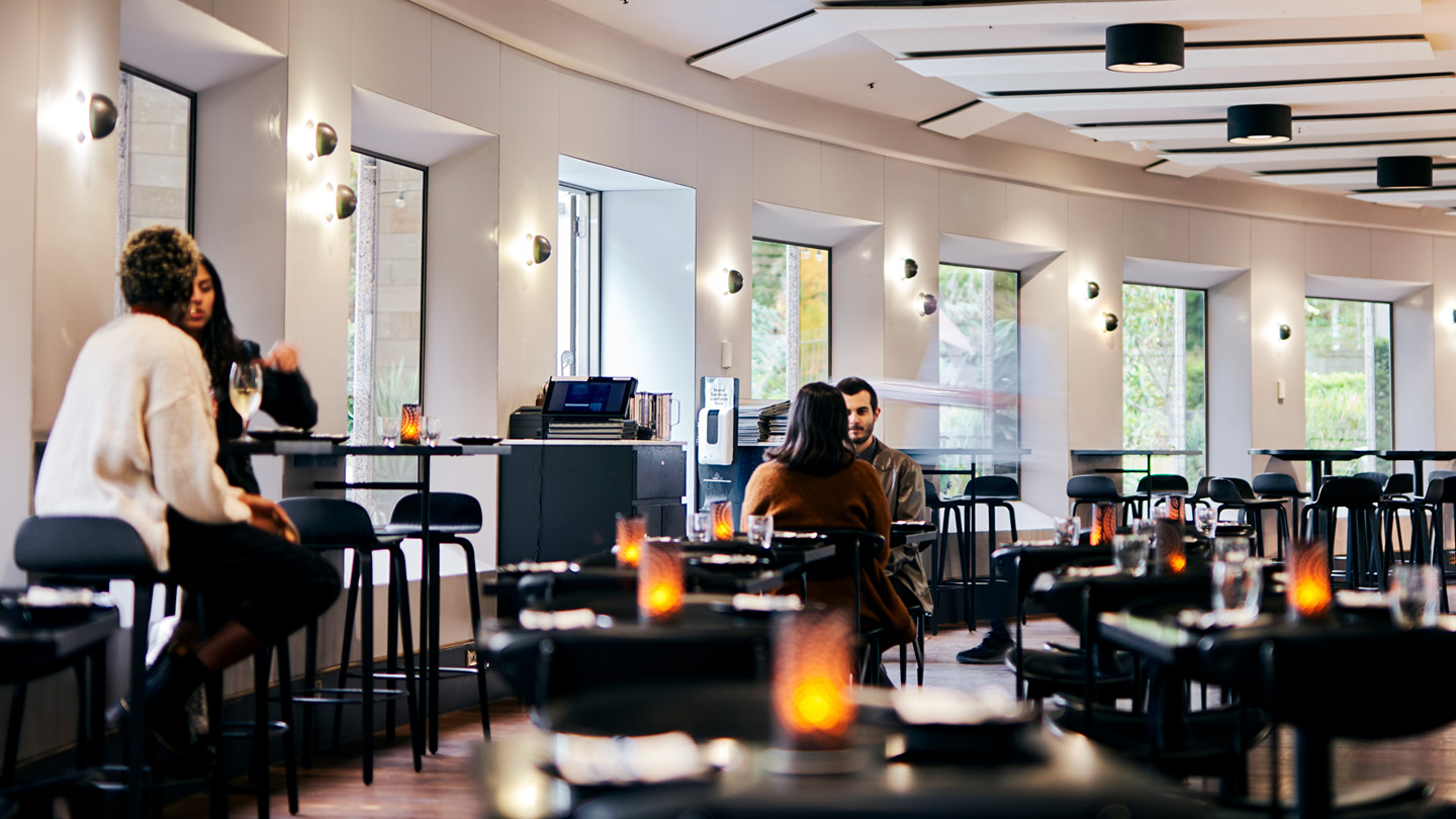 A image of the restaurant 'The Barre' during the day. Two customers are sitting on high bar stools to the left. In the middle of the restaurant is two other people sitting at a table. The bar furniture is all black, with lit candles on each table. 