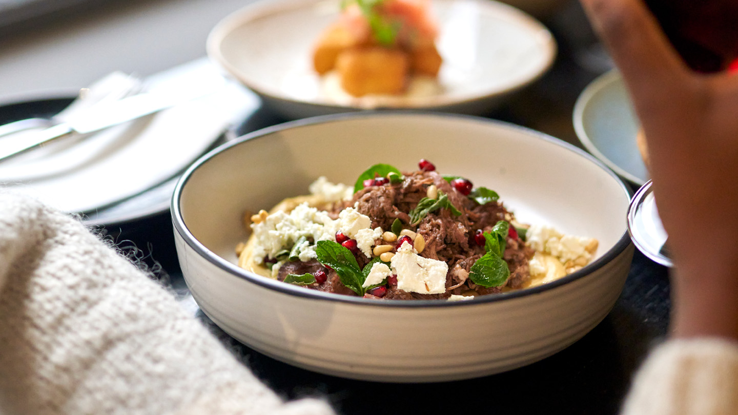 A meat dish in a round bowl. There is cheese, meat, pine buts, pomegranates and mint also in the bowl. 