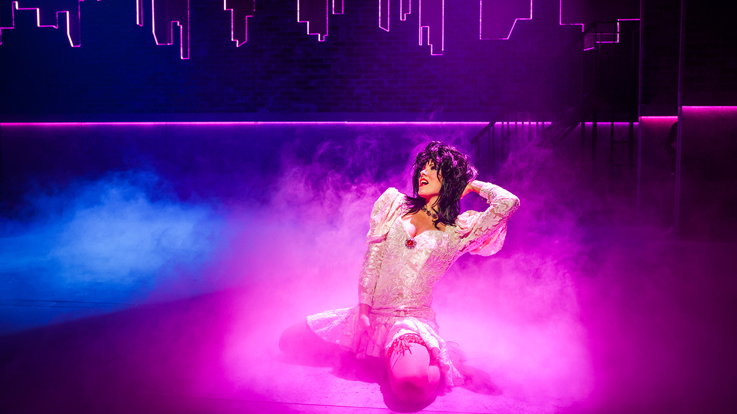 A production image from 'The Wedding Singer' depicting a woman with long, curly dark hair, dressed in a white dress and kneeling on the floor. One hand is behind her head, the other is near her crotch.