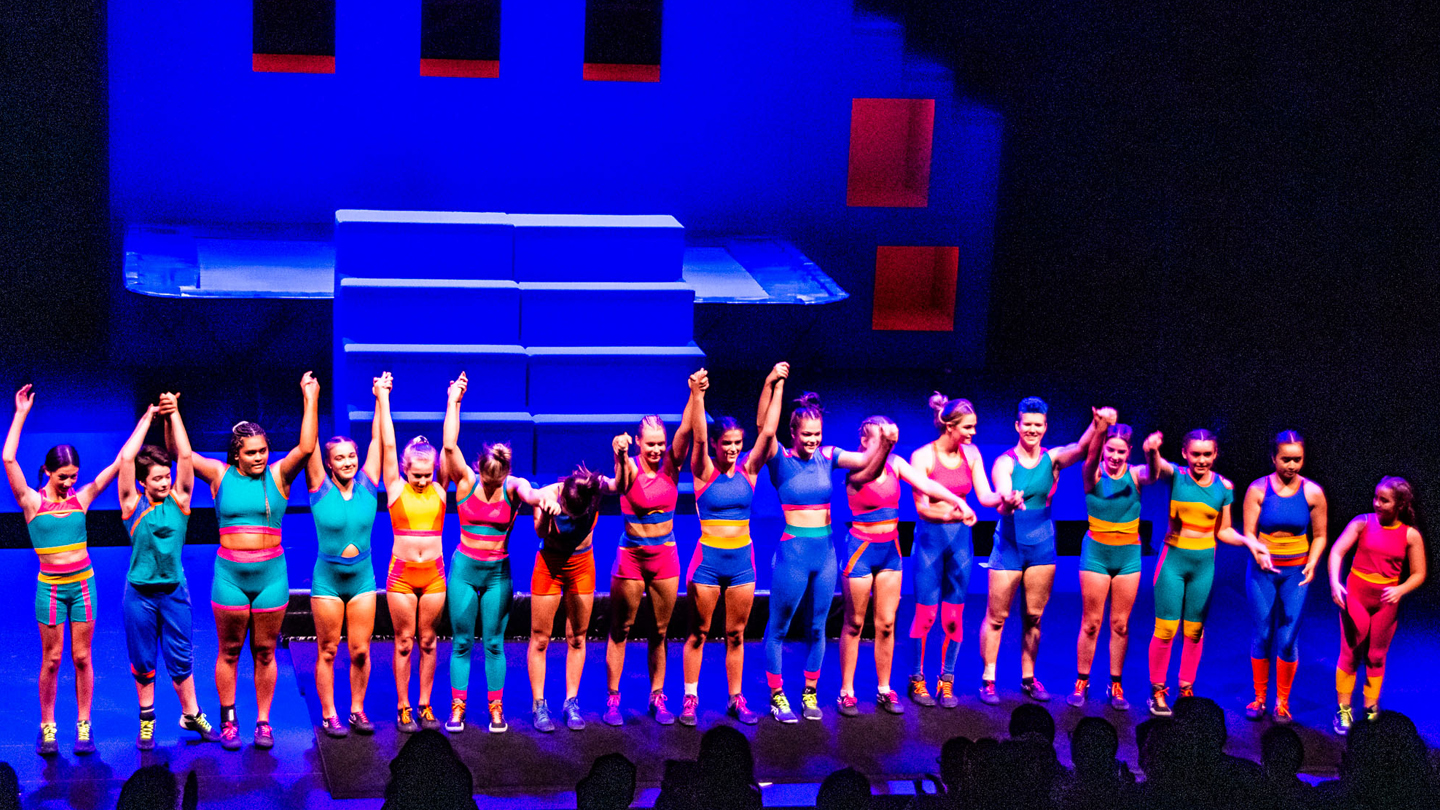 A production image from the circus performance 'Girls with Altitude'. A group of girls stand, holding hands, ready to bow to the audience. They are wearing colourful acrobatic costumes.