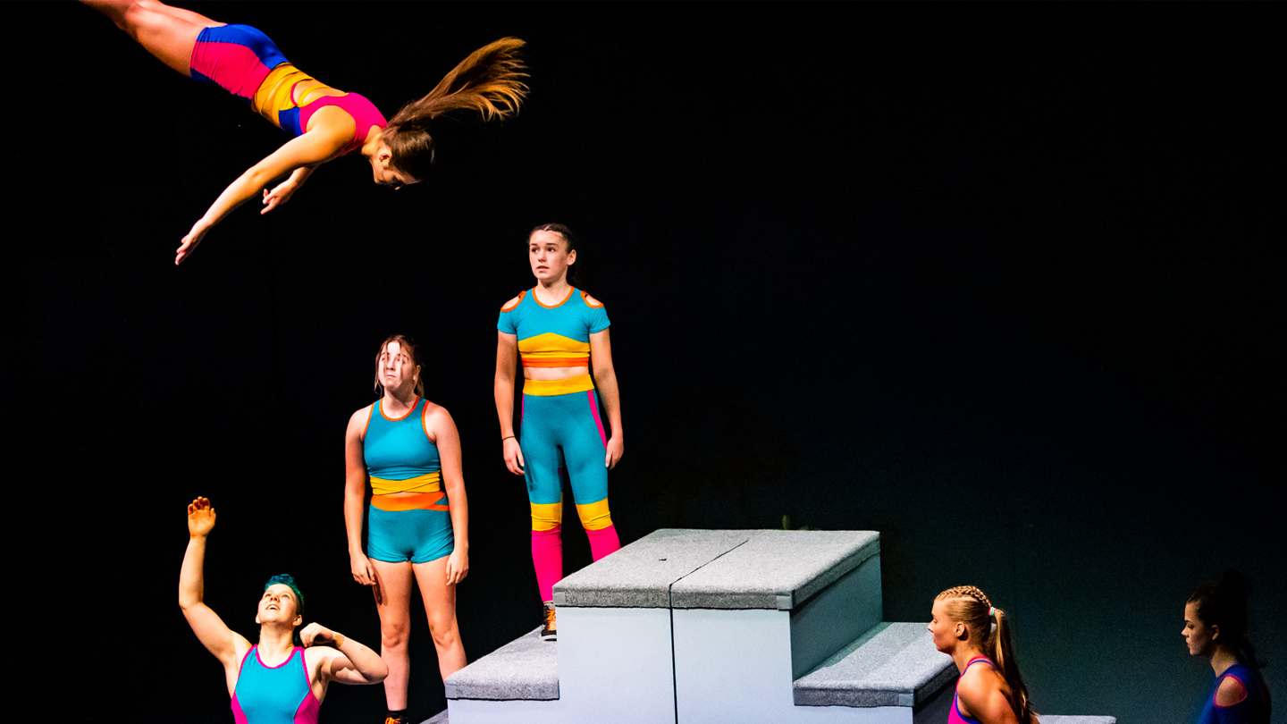A production image from the circus performance 'Girls with Altitude'. A group of girls stand, ready to catch another girl who is high in the air with arms extended out in front of her.