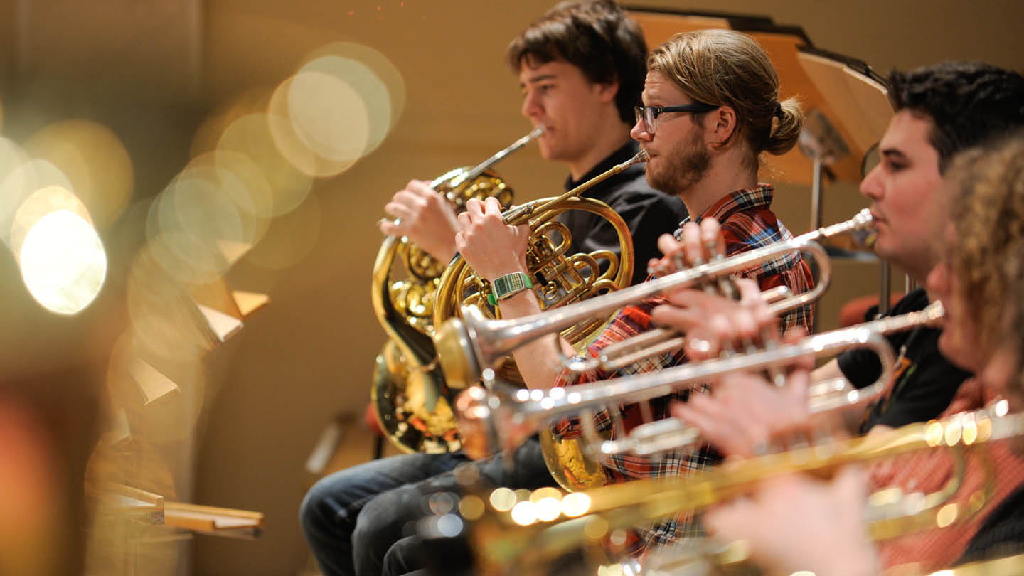 A close up of the brass section of Australian Youth Orchestra. Two men sit playing the french horn. To the right on them the row of trumpet players fades into the foreground.