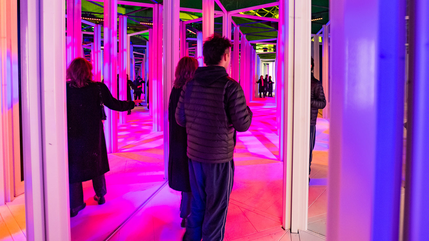 A female and male walking through a mirror maze with fluro lights around them.