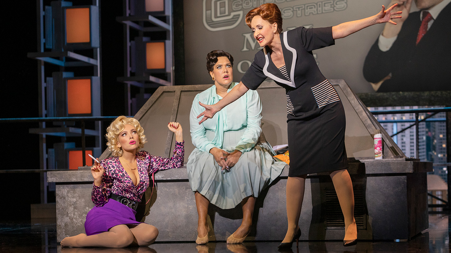 A production image from 9 TO 5 THE MUSICAL, showing the three main characters gathered together. On the left, the blond sits on the floor, her feet tucked to the side, in the middle, the brunette sits on a bench with her feet falling out to the side and a surprised expression, and on the right is a red head wearing a tight-fitting blazer and skirt, her hair done up in a chignon and her arms spread out to the sides, as she gestures widely and speaks.