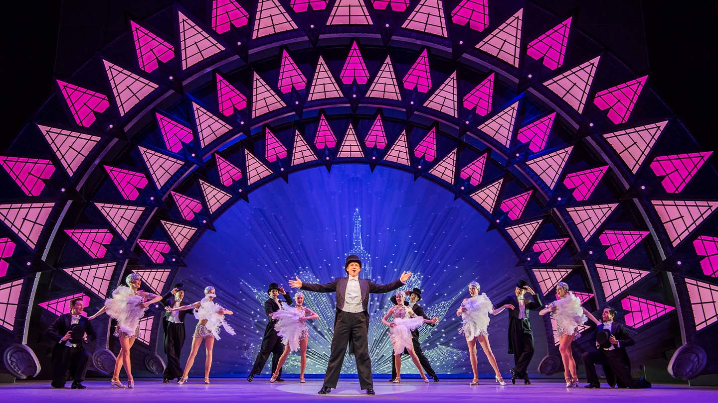 A man wearing a top hat and tails stands centre stage with his arms extended. Behind him are chorus girls in short silver dresses carrying large white fans. They're accompanied by well dressed men in suits.