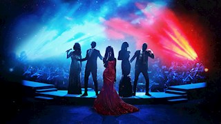 Five singers stand centre stage belting out a song. The two men wear sharp, black suits and the women are all wearing sequined ballgowns. Behind them is a full orchestra. There's vibrant red and blue lighting beaming across the stage and a haze of smoke.