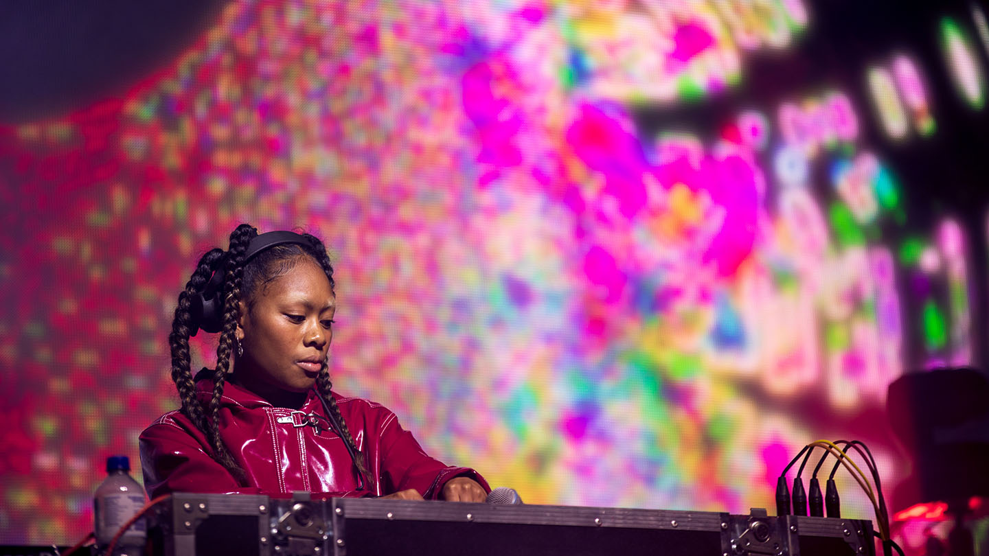 C.FRIM, a DJ with long dark braids wearing a shiny red jacket and headphones, performs onstage behind their decks. Behind them is a large video screen showing a psychedelic display of colours - green, purple, blue, green and yellow.