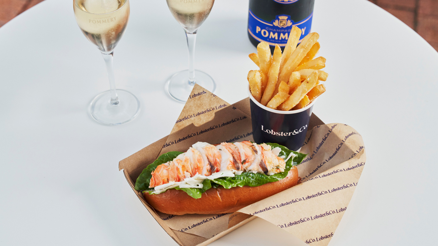 A close up of a lobster roll and a bucket of chips sitting on a cardboard food tray. Behind it are two flutes of champagne and a bottle of Pomery champagne. They're all sitting on a white table.