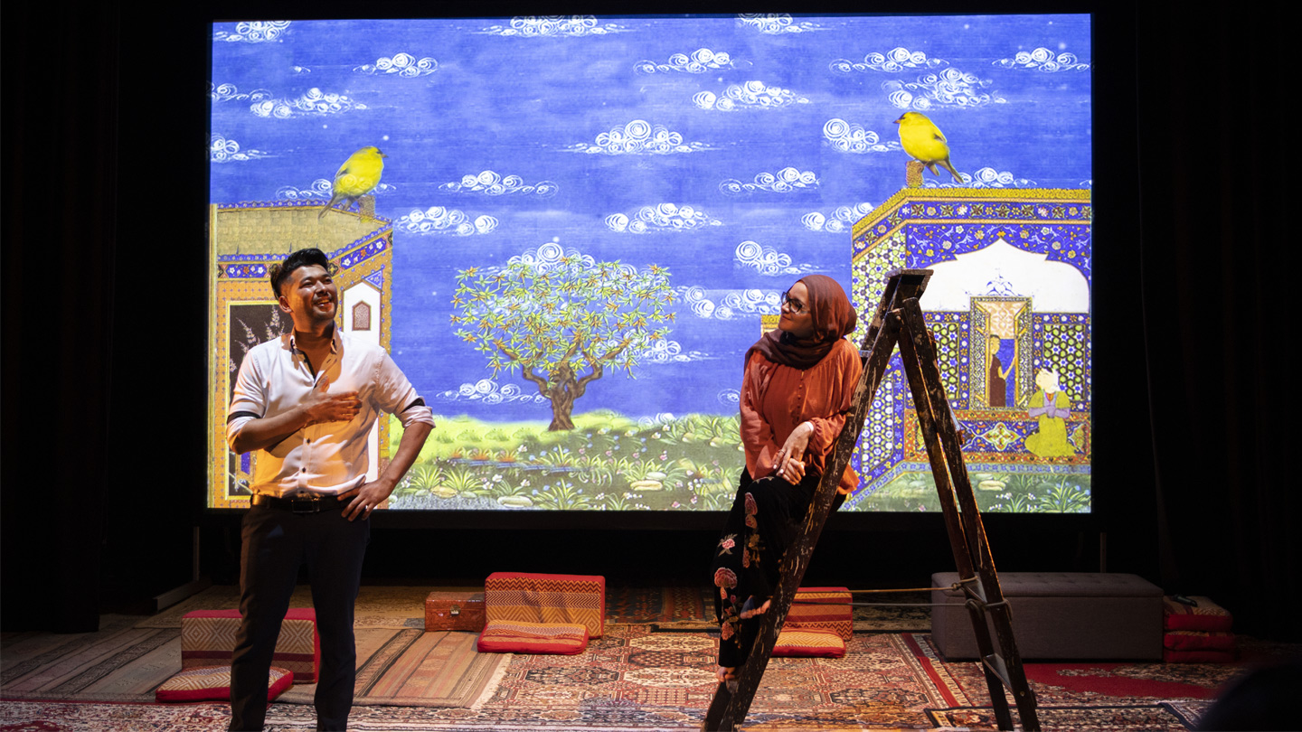 Production image from Dorr-e Dari. To the left a man in a white top and black trousers is talking, his feet apart, one hand on his hip and the other gesturing in front of his body. To the right a woman in niqab, wearing a red top and black trousers, is leaning with her whole body against one side of a wooden step ladder. Behind them is a large video display of an illustration showing a tree, two cliffs and a blue sky with white clouds.
