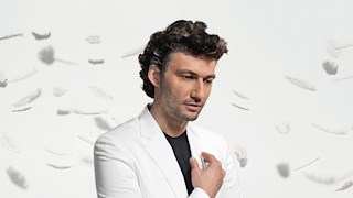 Jonas Kaufmann, a man with curly, dark hair, stands in a white space wearing a dark top and a white suit jacket. His hand is up to his heart and he is looking down with a peaceful expression. White feathers fall all around him.