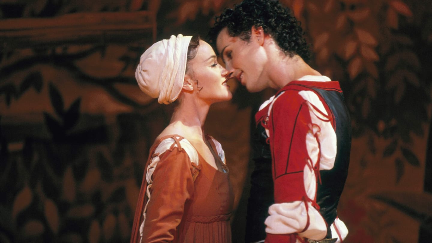 Two performers standing very close with heads leaning toward each other smiling and locking eyes. Female performer is in a white hat and mustard dress and male performers is in a red and black jacket with white shirt.