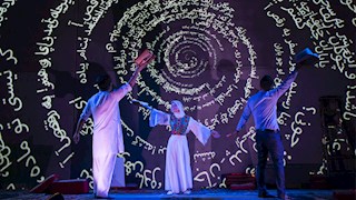 Three dimly lit figures wearing white dance onstage, their arms extended at angles from their bodies. They are lit with a slightly blue light from the left. Behind them is a large video display showing a massive spiral of Farsi script, shown as white text on a black background.