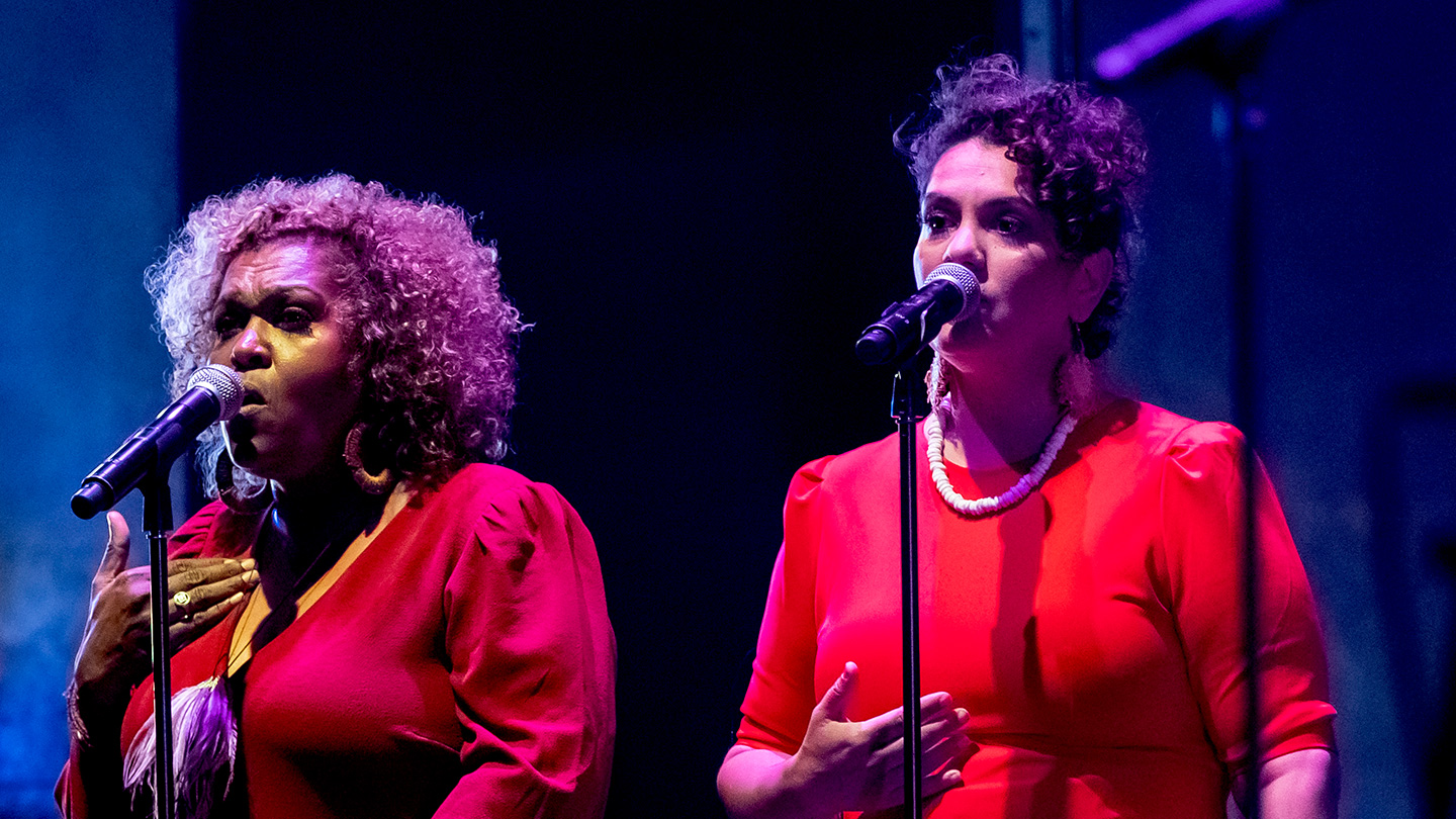 Emma Donovan and Deline Briscoe are singing into microphones on stage. They are both wearing red. Image credit: Tiffany Garvie.