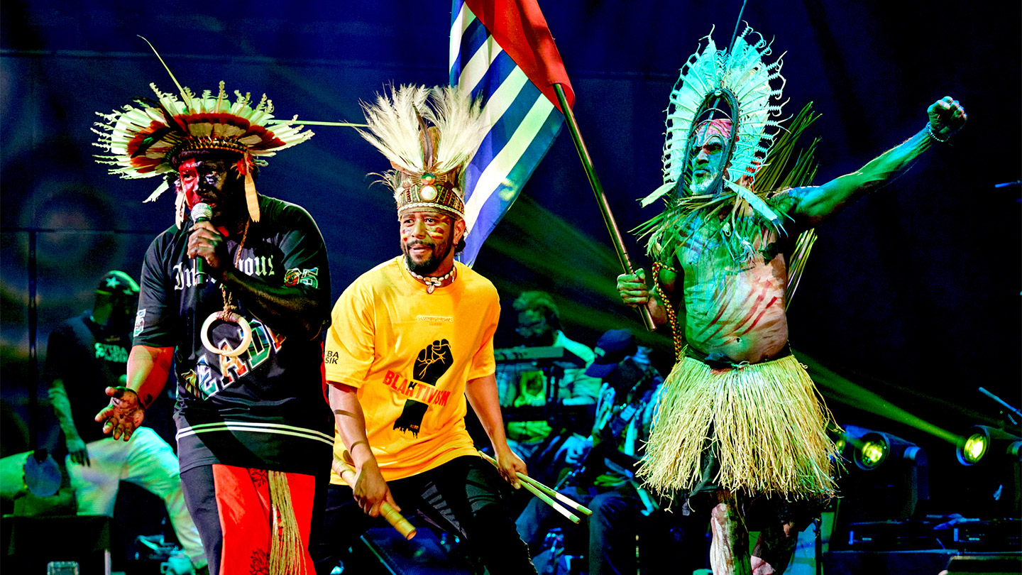 Sorong Samarai are performing on stage. They are wearing a mix of traditional dress and street clothes, they also have their faces painted. They hold a variety of instruments and behind them is a band. Image credit: James Taylor.