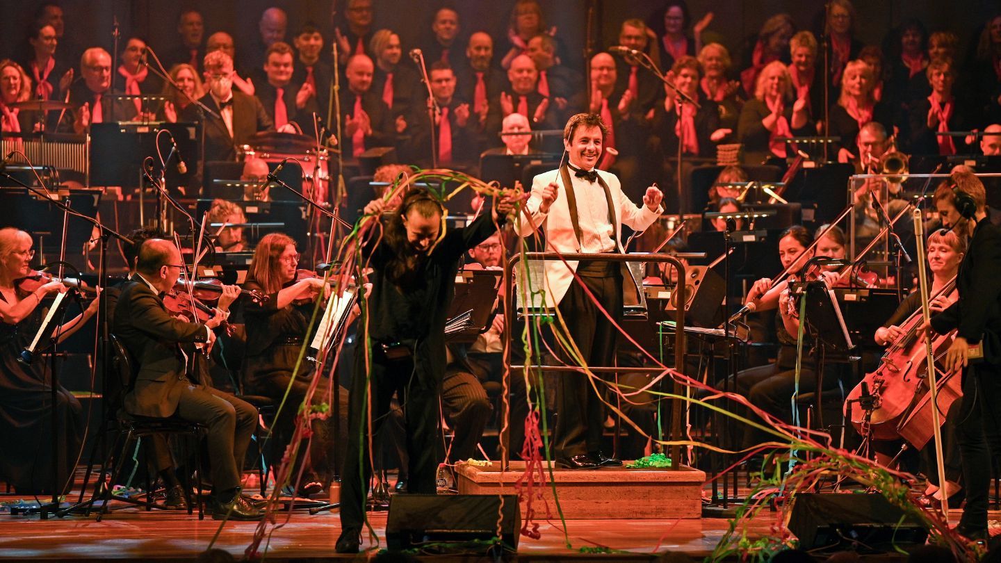 John Foreman conduction on stage in front of an orchestra. Streamers are being thrown onto the stage.
