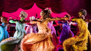 Cast of La Cage Aux Follies dancing on stage wearing bright coloured gowns.