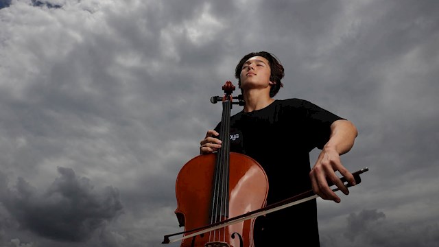 A teenage boy playing the cello. He is wearing a black shirt. The sky behind him is overcast.