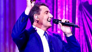 Daniel O'Donnell standing on stage singing into a microphone. he is wearing a blue suit and purple curtains are in the background.