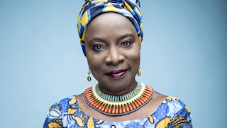 Angélique Kidjo  looking directly into the camera. She is wearing a matching blue and yellow top and head scarf. She is also wearing a colourful necklace and earrings. 