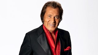Portrait of Engelbert Humperdinck. he is staring into the camera, he has brown hair and i swearing a black suit with a red shirt under. 