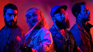 Hiatus Kaiyote band members Nai Palm, Perrin Moss, Paul Bender and Simon Mavin standing side by side, all looking a different direction with bright red and blue spotlights on them.