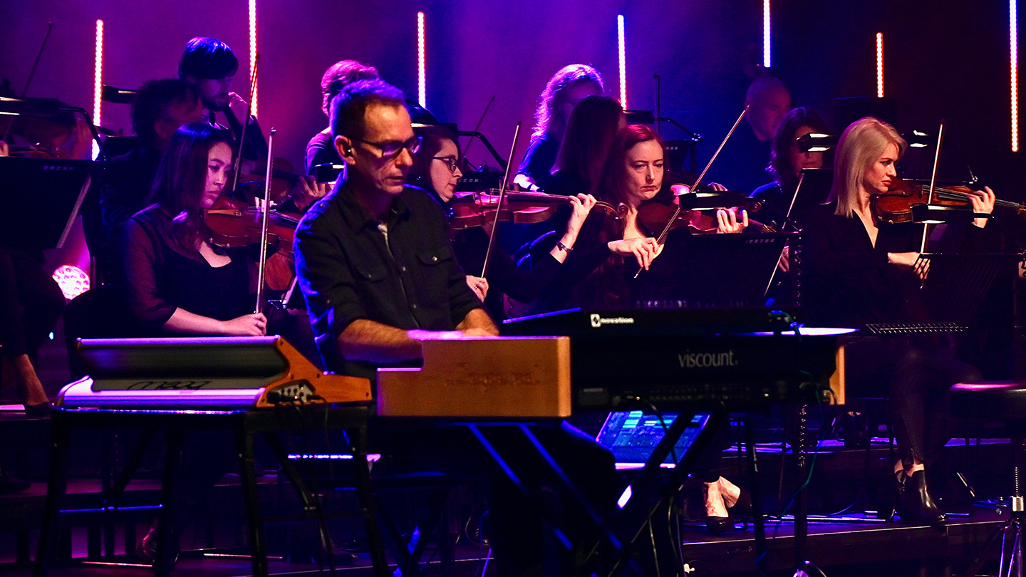 A man playing piano on stage. a string section sits behind him.