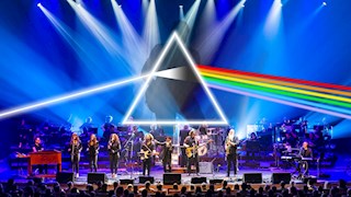 A large band on stage. Behind them is a projection of a man in silhouette, with an artwork of a light prism overlaid on top.