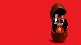 Brian Nankervis (he/him) Julia Zemiro (she/her) standing in a life size chocolate egg. The background is red.
