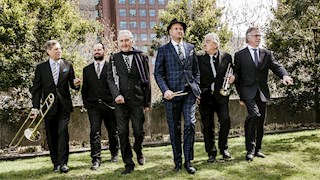Six members from The Syncopators wearing suits walking in a park with one holding a trumpet and one holding a trombone.