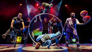 Image of 7 men on stage wearing all black. the man in the middle is balancing on his head, three men behind him are holding on to a large circle whilst another man jumps over it. to the left is a man on a bike and to the right is a man holding basketballs. 