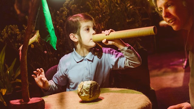A young girl pointing a cardboard tube into her mother's ear. They are sitting at a table with a garden in the background.