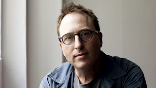 A headshot of Jon Ronson. He has brown hair, wears glasses and a blue shirt with a grey t-shirt underneath.