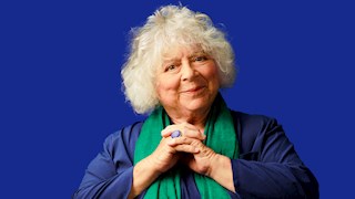 Miriam Margolyes smiling at the camera with her hands held together. She is wearing a blue top and a green scarf. The background is a matching blue colour to her shirt. 