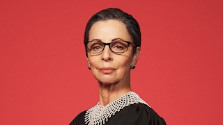 Heather Mitchell dressed as Ruth Bader Ginsburg is looking directly ahead. She wears glasses, a black top and small white beads around her neck. The background behind her is a pink-red.