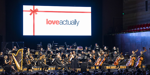 An Orchestra performing on stage with a large screen in the background with the words loveactually with a red ribbon.