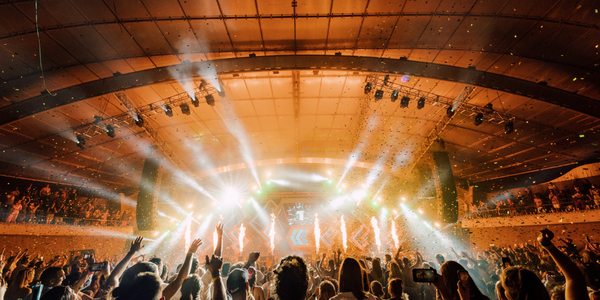 A wide shot of the Sidney Myer Music Bowl shell from behind with a packed crowd facing the stage under orange lights.