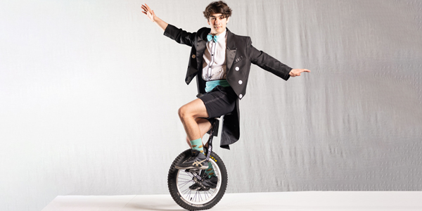 A boy riding a unicycle. He is wearing a black and white tuxedo with a light blue bow tie.