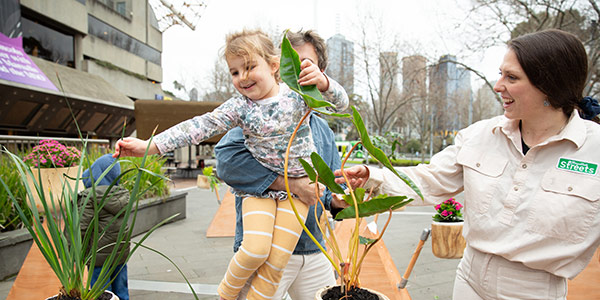 A child being lifted up by an adult. She is touching various plants sprouting out of wooden pots. The Arts Centre Melbourne Theatres building sits in the background.